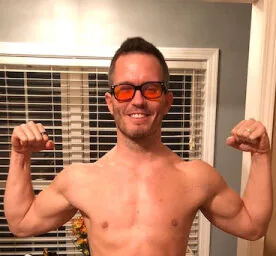 Smiling, shirtless male with sunglasses flexing biceps with arms upwards.